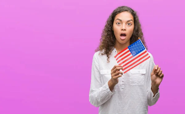Young hispanic woman holding flag of United Estates of America scared in shock with a surprise face, afraid and excited with fear expression