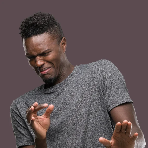 Young african american man wearing grey t-shirt disgusted expression, displeased and fearful doing disgust face because aversion reaction. With hands raised. Annoying concept.