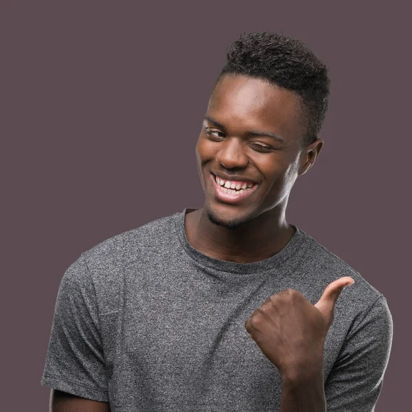 Young african american man wearing grey t-shirt smiling with happy face looking and pointing to the side with thumb up.