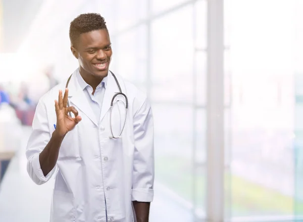 Young african american man wearing doctor coat doing ok sign with fingers, excellent symbol