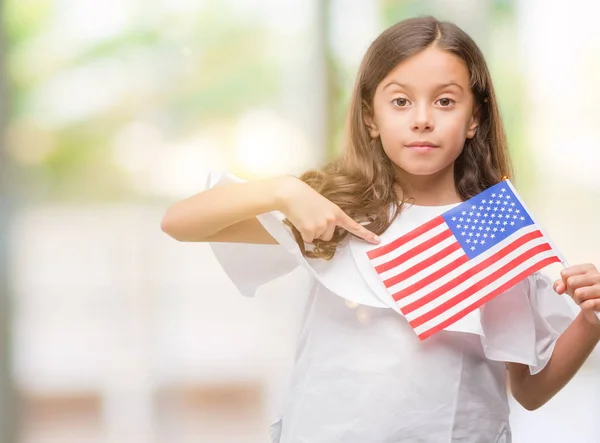 Brunette hispanic girl holding flag of United States of America with surprise face pointing finger to himself