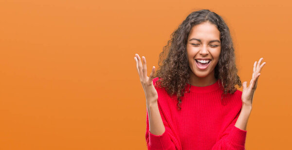 Young hispanic woman wearing red sweater celebrating mad and crazy for success with arms raised and closed eyes screaming excited. Winner concept