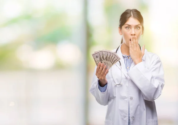 Young hispanic doctor woman holding dollars cover mouth with hand shocked with shame for mistake, expression of fear, scared in silence, secret concept