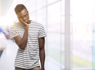Gözlük ve lacivert t-shirt düşünme seyir giyen genç Afro-Amerikan adam yorgun ve sıkılmış çapraz silah depresyon sorunları ile.