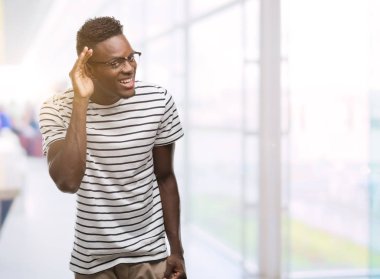 Genç Afro-Amerikan adam gözlük ve lacivert t-shirt ile gülümseyen bir işitme söylenti ya da dedikodu için dinleme kulak teslim. Sağırlık kavramı.