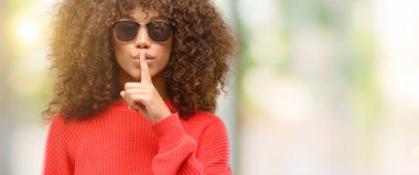 African american woman wearing sunglasses with index finger on lips, ask to be quiet. Silence and secret concept