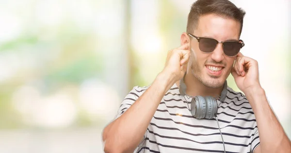 ハンサムな若い男が大声で音楽の音の腹が立つ式の指で耳を覆うヘッドフォンを身に着けています 聴覚障害者の概念 — ストック写真