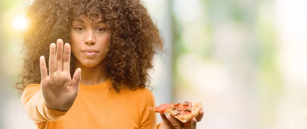 African american woman with a pizza slice with open hand doing stop sign with serious and confident expression, defense gesture