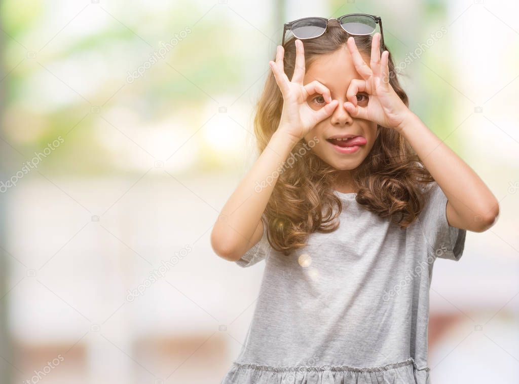 Brunette hispanic girl wearing sunglasses doing ok gesture like binoculars sticking tongue out, eyes looking through fingers. Crazy expression.