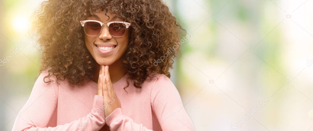 African american woman wearing pink sunglasses praying with hands together asking for forgiveness smiling confident.