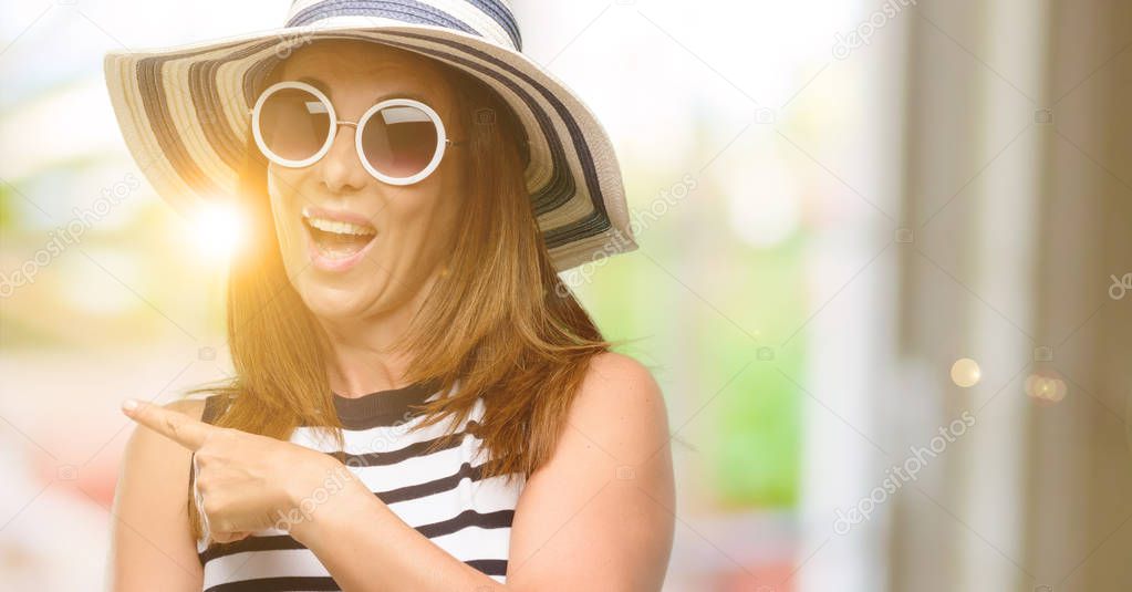Middle age cool woman wearing summer hat and sunglasses pointing away side with finger