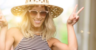 Genç kadın parmakları tong ve verme zafer işaretiyle gösterilen kamera bakarak yaz şapka güneş gözlüğü kullanarak