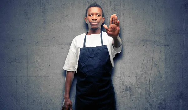 African man shop owner wearing apron annoyed with bad attitude making stop sign with hand, saying no, expressing security, defense or restriction, maybe pushing