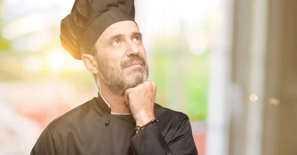 Senior cook man, wearing chef hat thinking and looking up expressing doubt and wonder