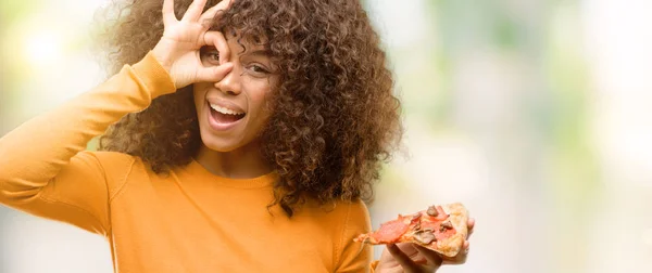 African american woman with a pizza slice with happy face smiling doing ok sign with hand on eye looking through fingers