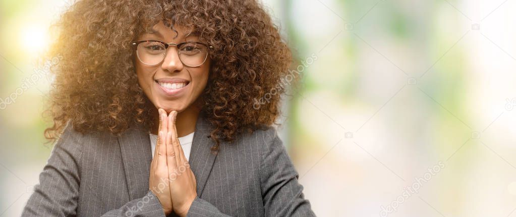 African american business woman wearing glasses praying with hands together asking for forgiveness smiling confident.