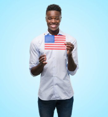 ABD bayrağı ile ayakta ve diş gösterilen kendine güvenen bir gülümsemeyle gülümseyerek mutlu bir yüz tutan genç Afro-Amerikan adam