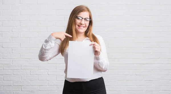 Young adult business woman standing over white brick wall holding blank paper with surprise face pointing finger to himself