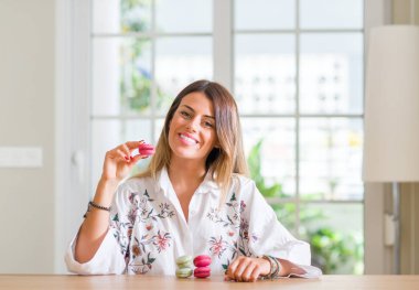 Genç kadının evde duran ve diş gösterilen kendine güvenen bir gülümsemeyle gülümseyerek mutlu bir yüz ile macarons yediğini