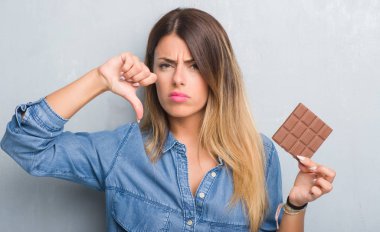 Young adult woman over grey grunge wall eating chocolate bar with angry face, negative sign showing dislike with thumbs down, rejection concept