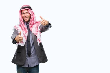 Genç yakışıklı Arap adam uzun saçlı keffiyeh el ile pozitif jest yapıyor onaylama izole arka plan üzerinde giyen başparmak gülümseyen ve başarı için mutlu. Kamera, kazanan jest arıyorsunuz.