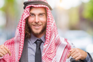 Uzun saçlı keffiyeh yüzünde gülümseme ile kendinden emin görünüyorsun, kendini gururlu ve mutlu parmakları ile işaret izole arka plan üzerinde giyen genç Arap yakışıklı.
