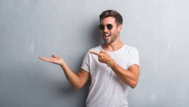 Hayret ve el ile sunulması ve parmak ile işaret ederken kameraya gülümseyen güneş gözlüğü takıyor gri grunge duvarın üzerinden yakışıklı genç.