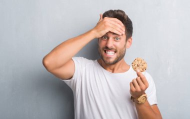 Handsome young man over grey grunge wall eating chocolate chip cooky stressed with hand on head, shocked with shame and surprise face, angry and frustrated. Fear and upset for mistake. clipart