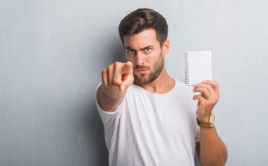 Kamera ve size parmak ile işaret boş defter tutan gri grunge duvarın üzerinden yakışıklı genç adam, el işareti, pozitif ve kendinden emin bir jest ön