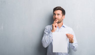 Yakışıklı genç gri grunge duvar holding boş kağıt sayfası sözleşme ciddi bir yüz düşünme hakkında soru, çok karışık bir fikir üzerinde