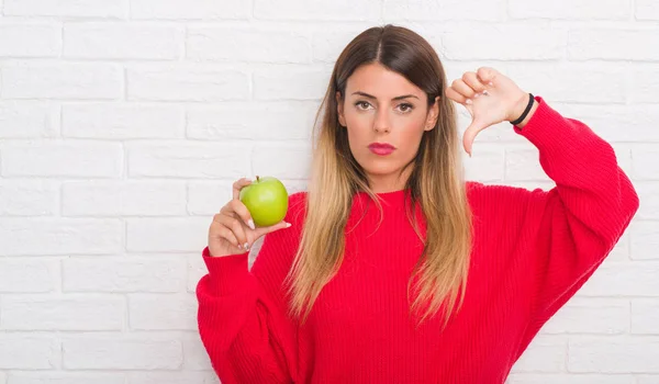 年轻的成年妇女在白色砖墙吃新鲜绿色苹果与愤怒的面孔 消极标志显示不喜欢与拇指下来 拒绝概念 — 图库照片