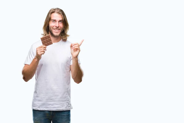 长头发的年轻英俊的人吃巧克力酒吧在隔绝的背景非常愉快指向用手和手指到边 — 图库照片
