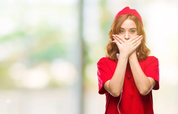 ヘッドフォンとショックのミスのための手で口を覆っているキャップを身に着けている孤立した背景に若い美しい流行に敏感な女性 秘密の概念 — ストック写真