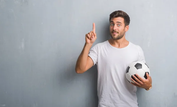 幸せそうな顔 ナンバーワンのアイデアや質問ポインティング指で驚いたサッカー サッカー ボールを保持している灰色のグランジの壁を越えてハンサムな若い男 — ストック写真