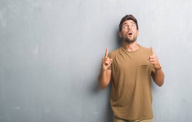 Yakışıklı genç gri grunge üzerinden hayran ve sürpriz ve parmakları ile işaret duvar ve silah kaldırdı.