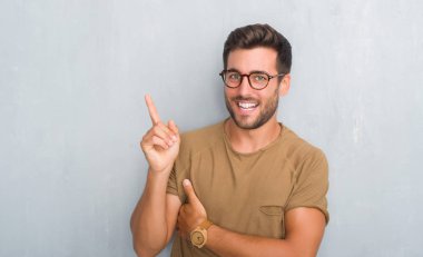 Kameraya bakarak yan el ve parmak ile işaret eden yakışıklı genç adam gözlüklü kocaman bir gülümseme ile yüzünde, gri grunge duvarın üzerinden.