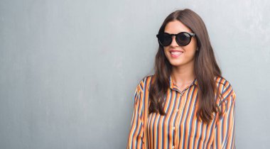 Genç esmer kadın ayakta ve diş gösterilen kendine güvenen bir gülümsemeyle gülümseyerek mutlu bir yüz ile büyük güneş gözlüğü takıyor grunge gri duvarın üzerinden