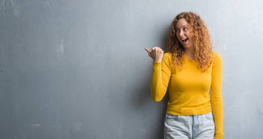 Genç Kızıl saçlı kadın işaret ve başparmak kadar mutlu yüz gülümseyen tarafı ile gösterilen gri grunge duvarın üzerinden