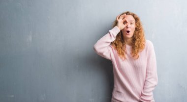 Sürpriz yüz, göz parmaklarının arasından seyir ile genç redhead gri grunge duvar giyen üzerinde pembe kadın kazak ok jest yapıyor şok. İmansız ifade.
