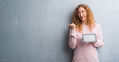 İşaret ve başparmak kadar mutlu yüz gülümseyen tarafı ile gösterilen tablet kullanarak gri grunge duvar genç Kızıl saçlı kadın