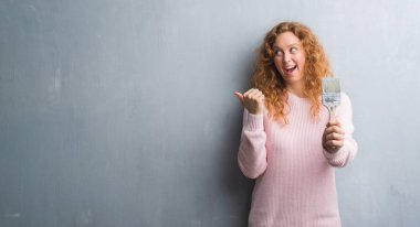 Gri grunge duvar işaret ve başparmak kadar mutlu yüz gülümseyen tarafı ile gösterilen boya fırçası tutan genç Kızıl saçlı kadın