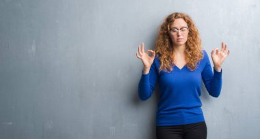 Genç Kızıl saçlı kadın gri grunge duvarın üzerinden rahatla ve gülümseyen gözlerle kapalı parmak jestle meditasyon yapıyor. Yoga kavramı.