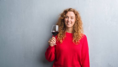 Genç Kızıl saçlı kadın ayakta ve diş gösterilen kendine güvenen bir gülümsemeyle gülümseyerek mutlu bir yüz ile bir kadeh şarap içmek gri grunge duvarın üzerinden