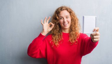 Gri grunge duvar gösteren boş defter parmak, mükemmel sembolü ile ok işareti yaparken genç Kızıl saçlı kadın