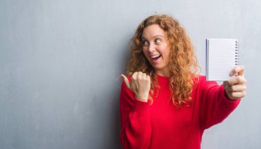 İşaret ve başparmak kadar mutlu yüz gülümseyen tarafı ile gösterilen gri grunge duvar gösteren boş defter genç Kızıl saçlı kadın