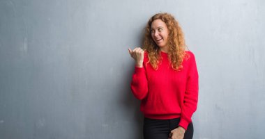 Seyir ve başparmak yanında yukarıyı mutlu yüz ile gülümseyen genç redhead gri grunge duvar giyen üzerinde kırmızı kadın kazak.