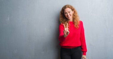 Genç redhead gri grunge duvar giyen üzerinde kırmızı kadın kazak zafer işareti yapıyor parmak gösterilen kamera seyir gülümseyerek. İki numaralı.
