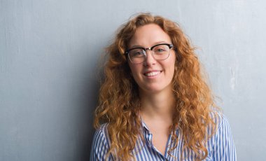 Gri grunge duvar ayakta ve diş gösterilen kendine güvenen bir gülümsemeyle gülümseyerek mutlu bir yüz ile genç Kızıl saçlı kadın