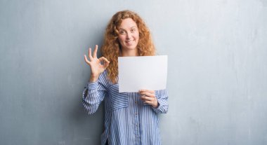 Gri grunge duvar parmak, mükemmel sembolü ile ok işareti yapıyor boş kağıt sayfası tutan genç Kızıl saçlı kadın