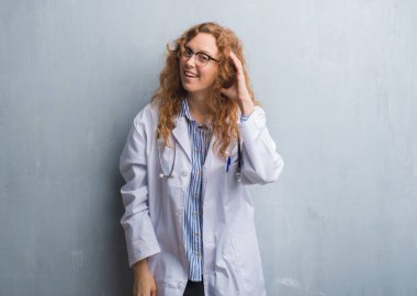 Genç redhead doktor kadın gri grunge duvar bir işitme söylenti ya da dedikodu için dinleme kulak üzerinde el ile gülümseyen bir palto giyiyor. Sağırlık kavramı.